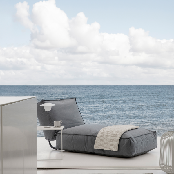 Modern grey lounge chair by the ocean, featuring a side table with a lamp and cup.
