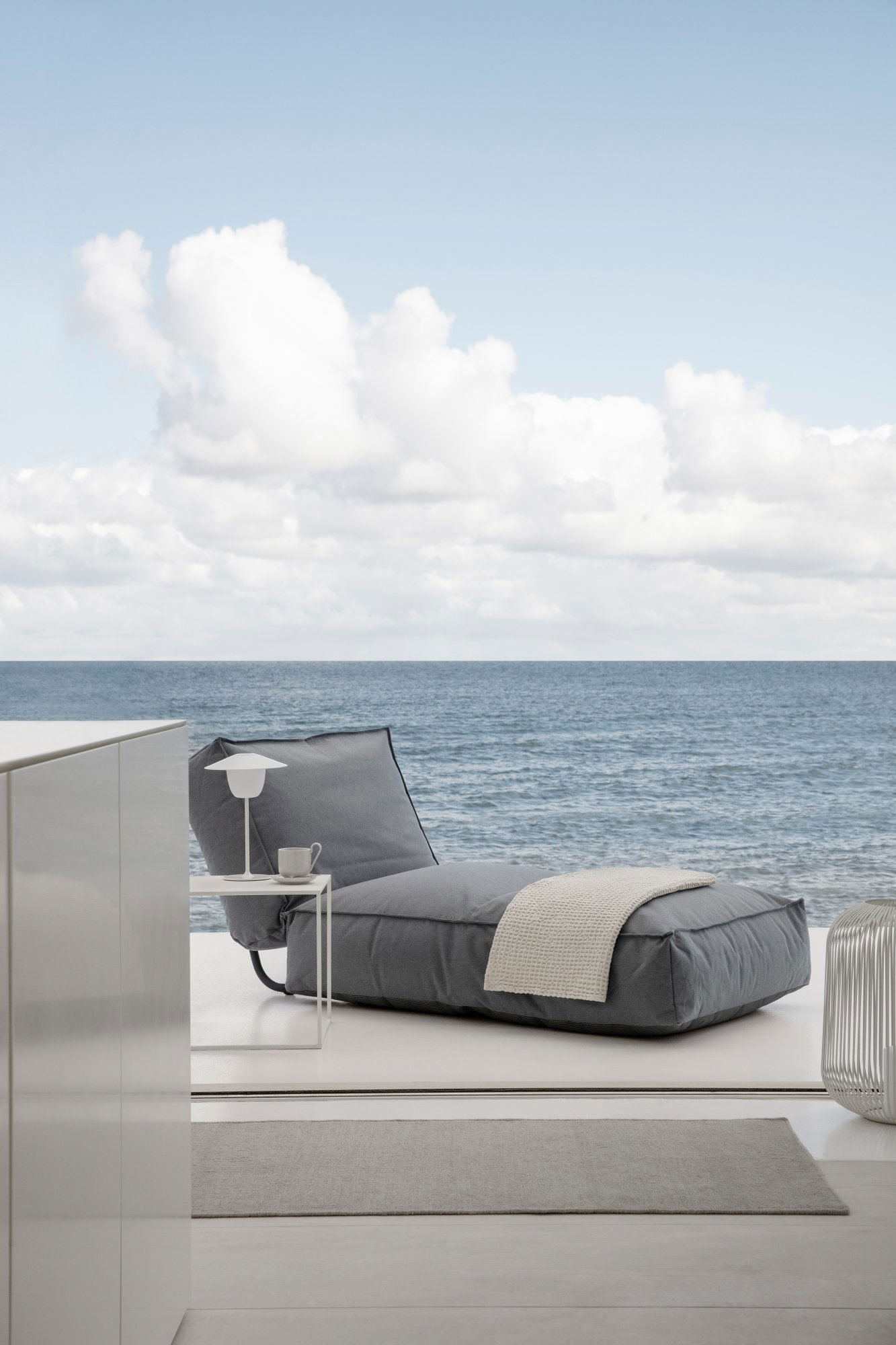 Modern grey lounge chair by the ocean, featuring a side table with a lamp and cup.