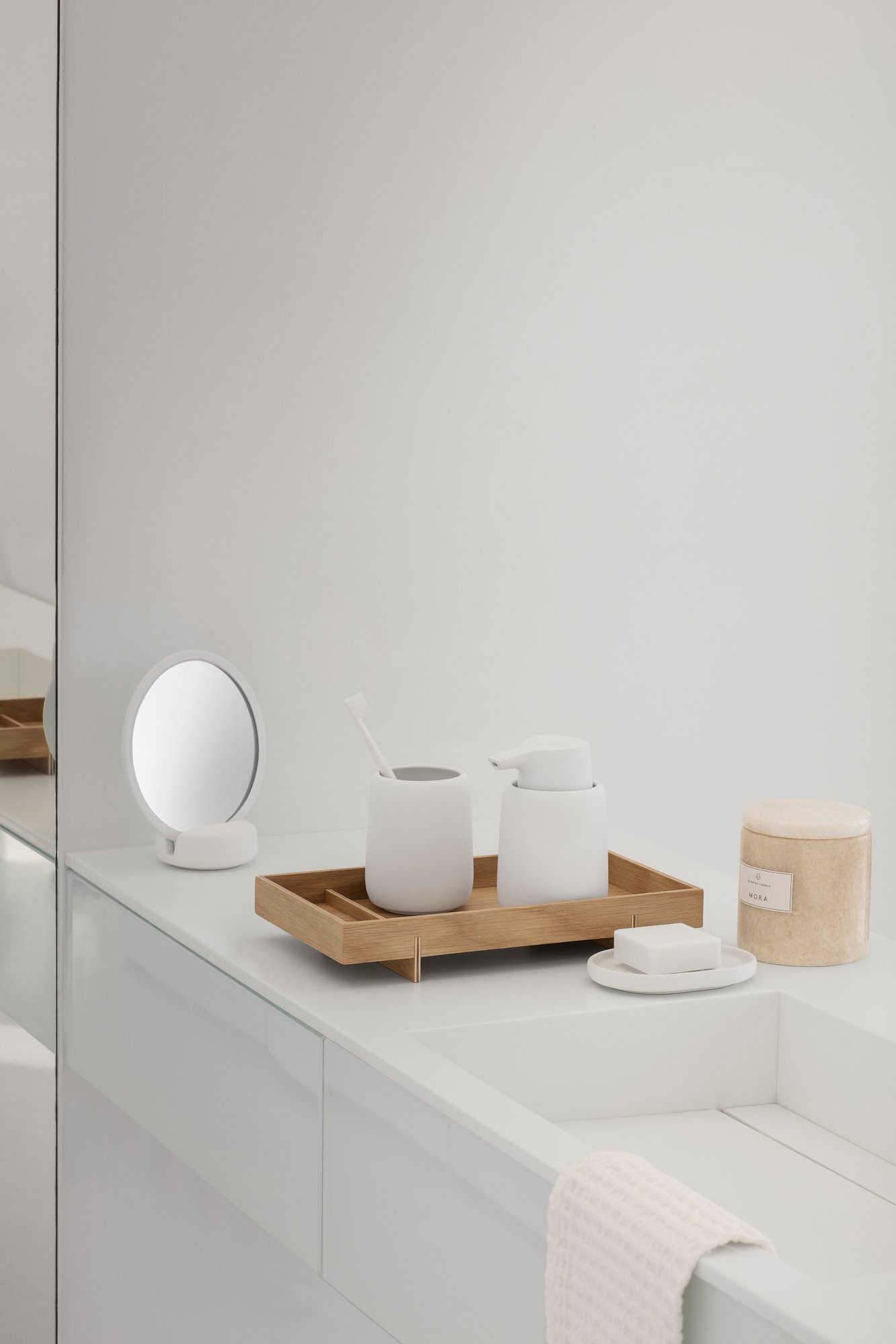 Minimalist bathroom decor featuring wooden tray, white containers, and a round mirror.