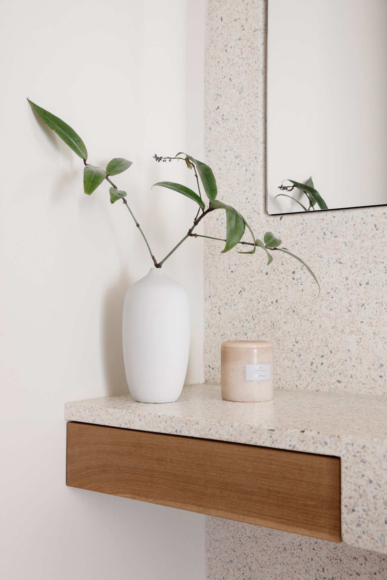 Minimalistische Dekoration mit einer weißen Vase, die eine Pflanze neben einer Kerze auf einer strukturierten Oberfläche zeigt.