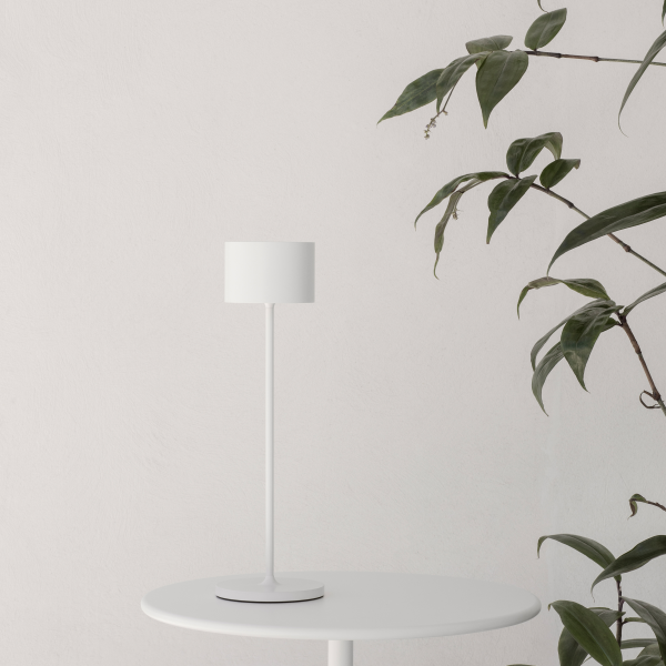 Minimalistische weiße Lampe auf einem Tisch neben einer grünen Pflanze vor einem hellen Wandhintergrund.
