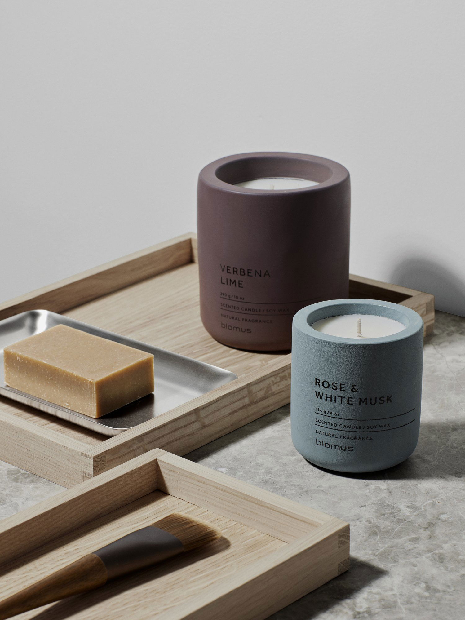Scented candles in modern holders next to a soap bar on a wooden tray.