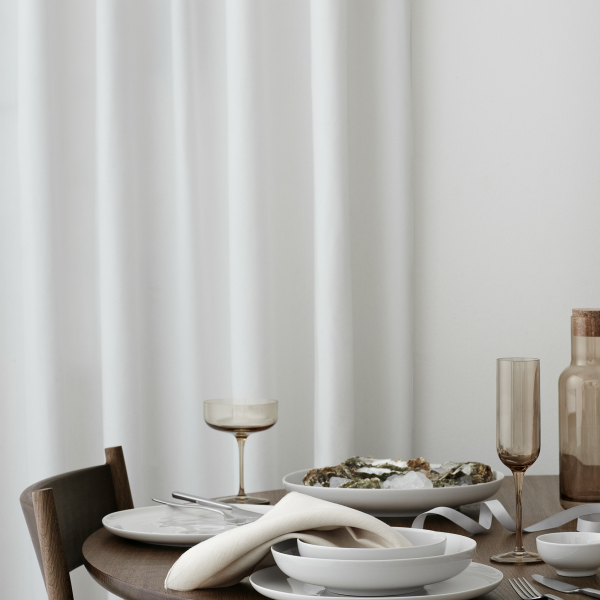 Elegantes Esstischset mit weißen Tellern, Gläsern und einer Platte mit Essen unter sanften Vorhängen.