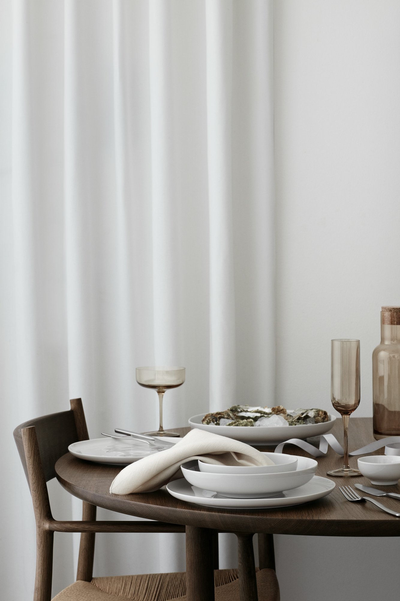 Elegantes Esstischset mit weißen Tellern, Gläsern und einer Platte mit Essen unter sanften Vorhängen.