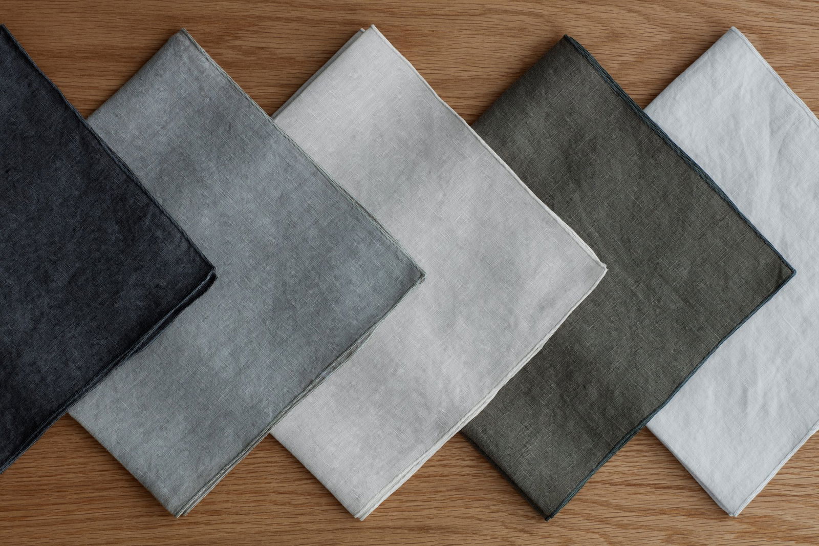 Assorted linen napkins in gray, white, and olive green arranged on a wooden surface.