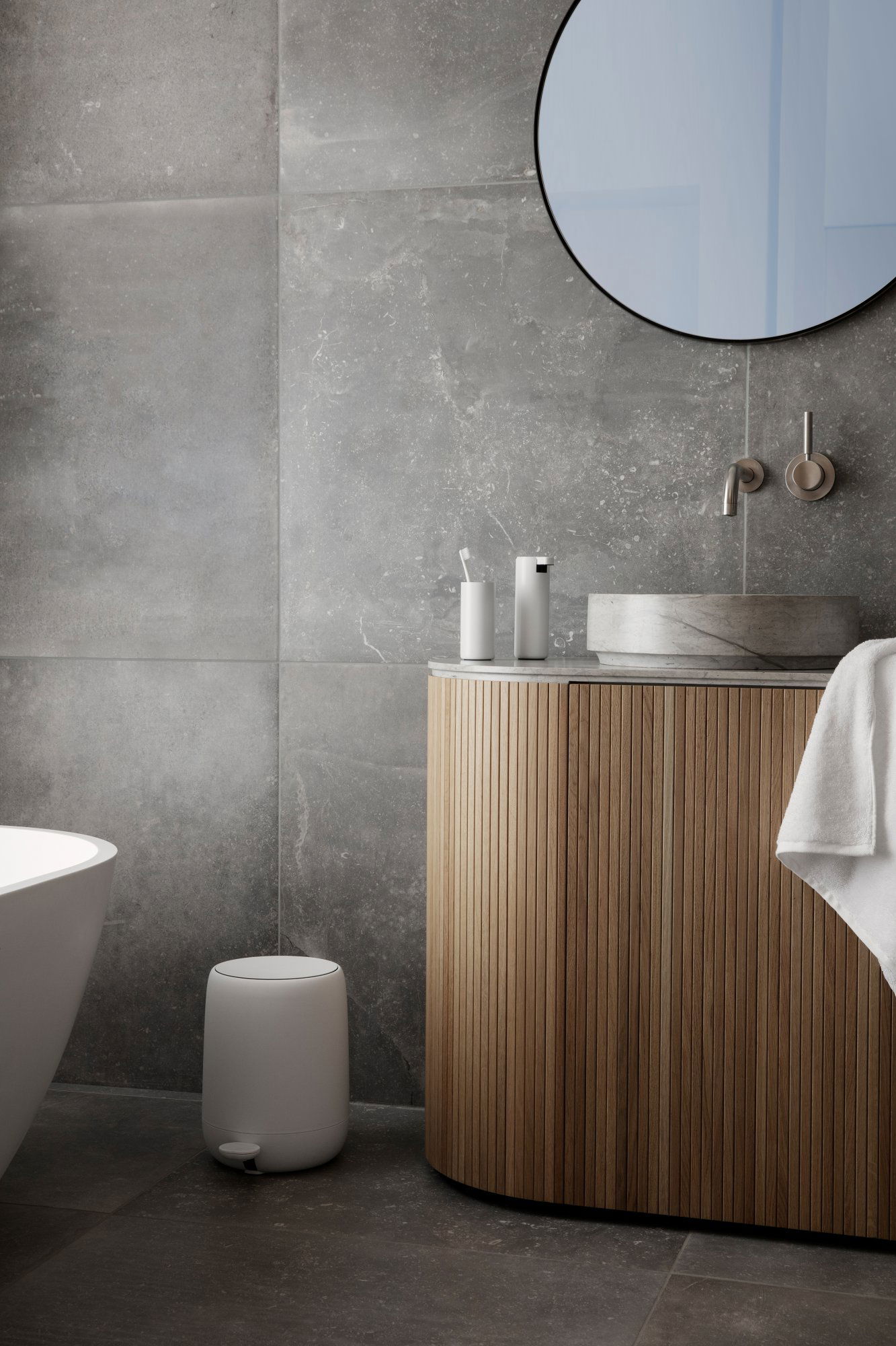 Modern bathroom with gray tiles, wooden vanity, circular mirror, and minimalist decor.