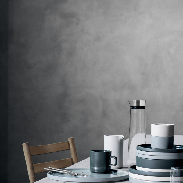Modernes Ess-Setup mit minimalistischem Geschirr, darunter Tassen und Behälter, vor einem strukturierten Hintergrund.