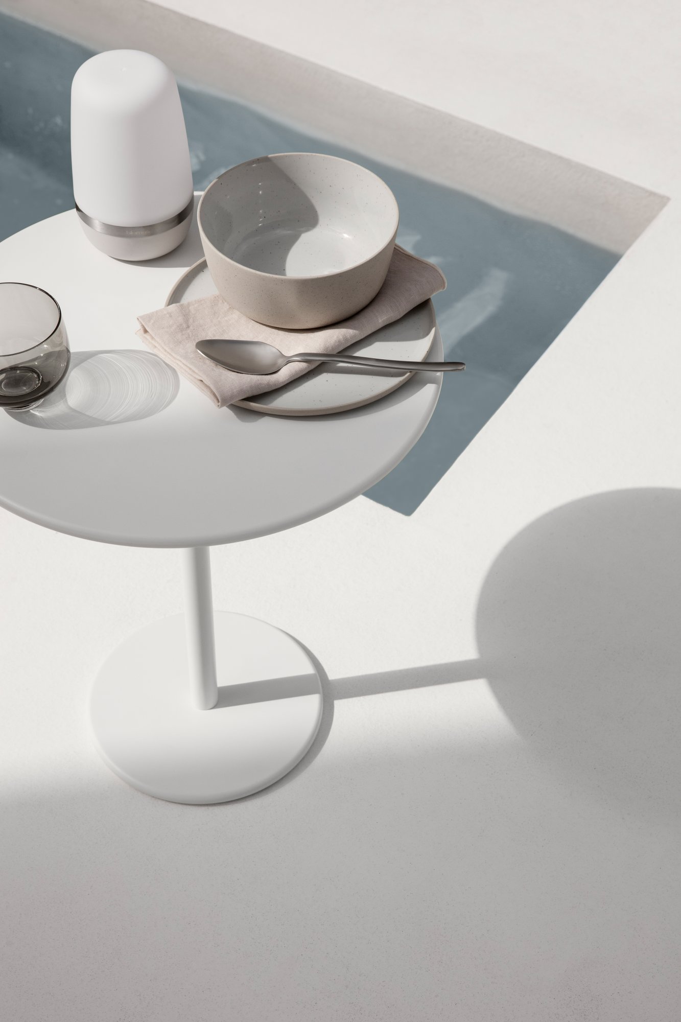 Minimalistisches Esssetup auf einem weißen Tisch mit Tellern und Gläsern neben einem Pool.