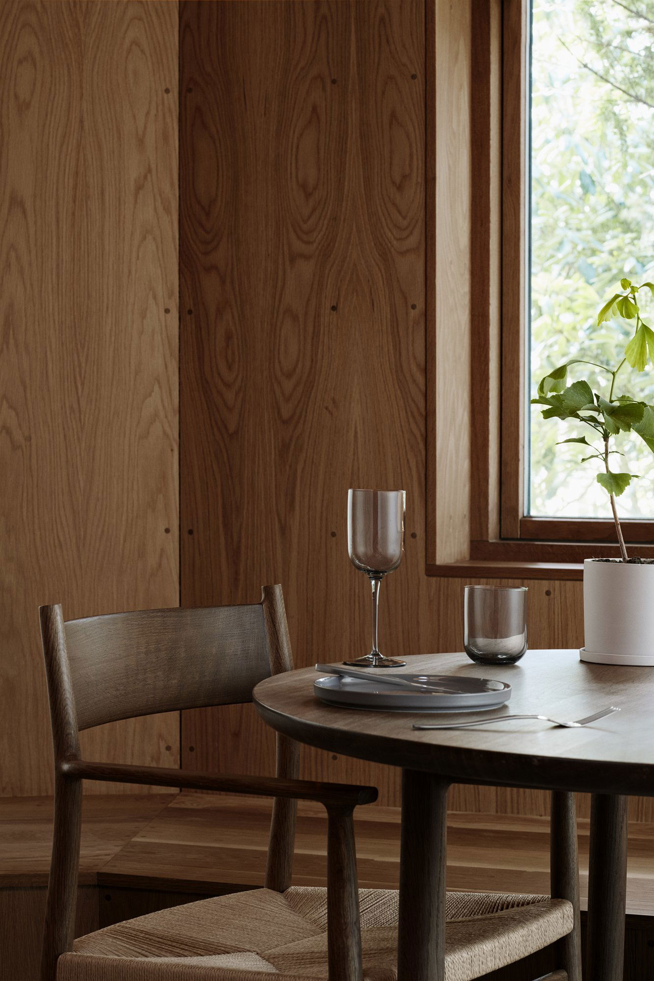 Modern dining setting with wooden walls, a table, glasses, and a potted plant by the window.