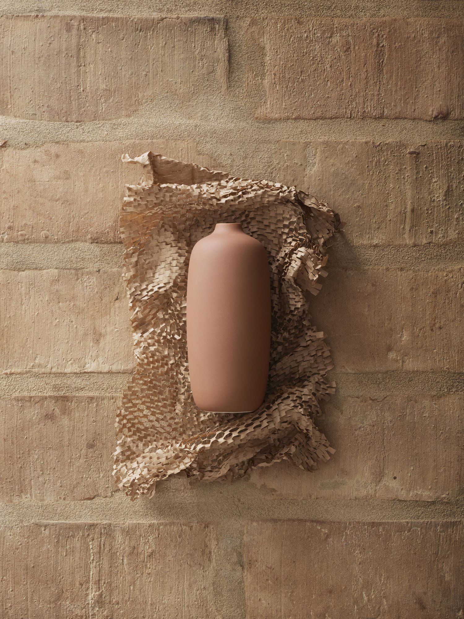 Minimalist brown vase on textured paper against a rustic brick wall background.