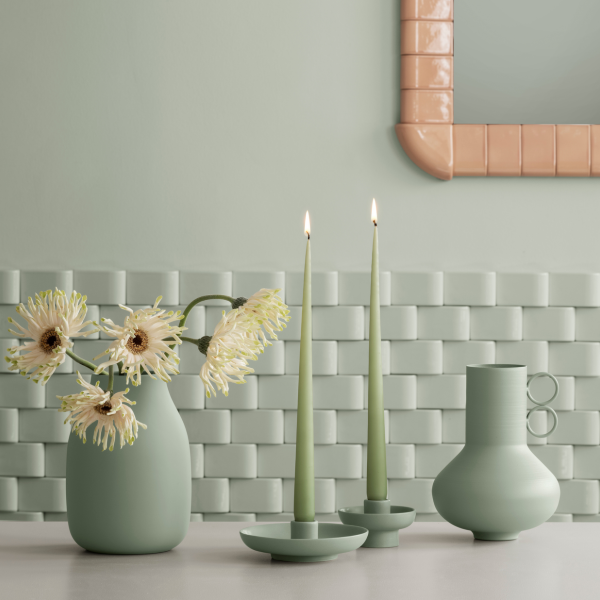 Elegante grüne Dekoration mit Vasen, Kerzen und Blumen vor einem strukturierten Wandhintergrund.