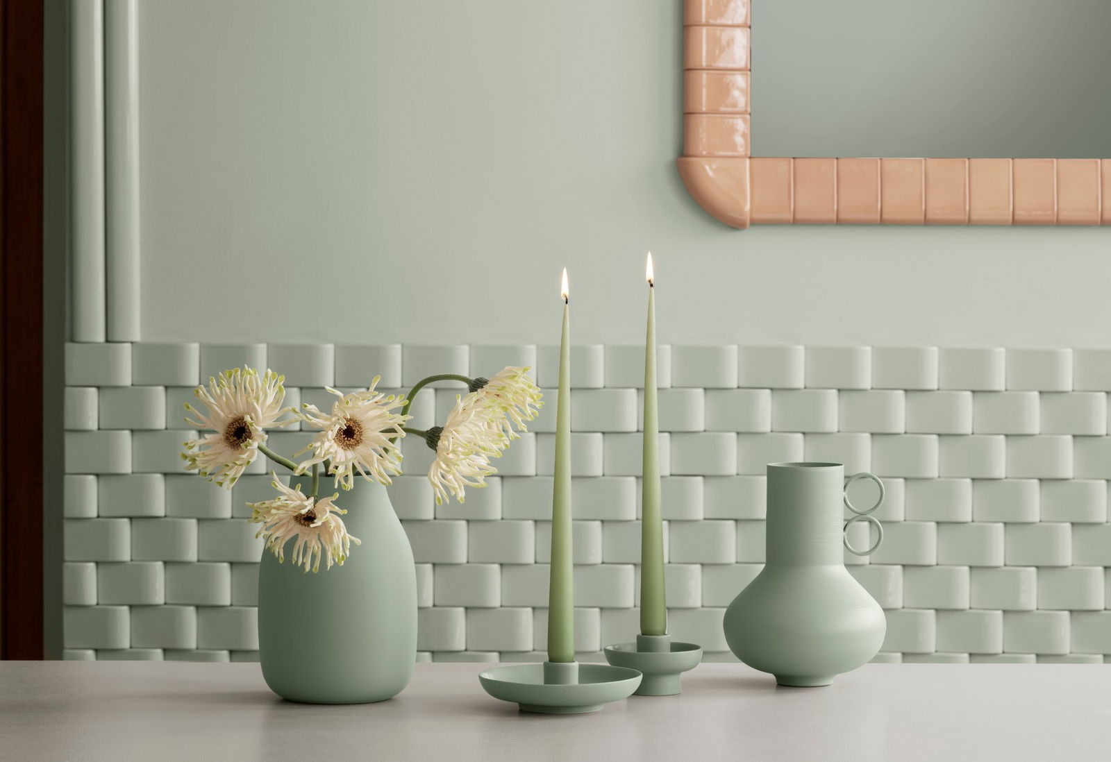 Elegante grüne Dekoration mit Vasen, Kerzen und Blumen vor einem strukturierten Wandhintergrund.