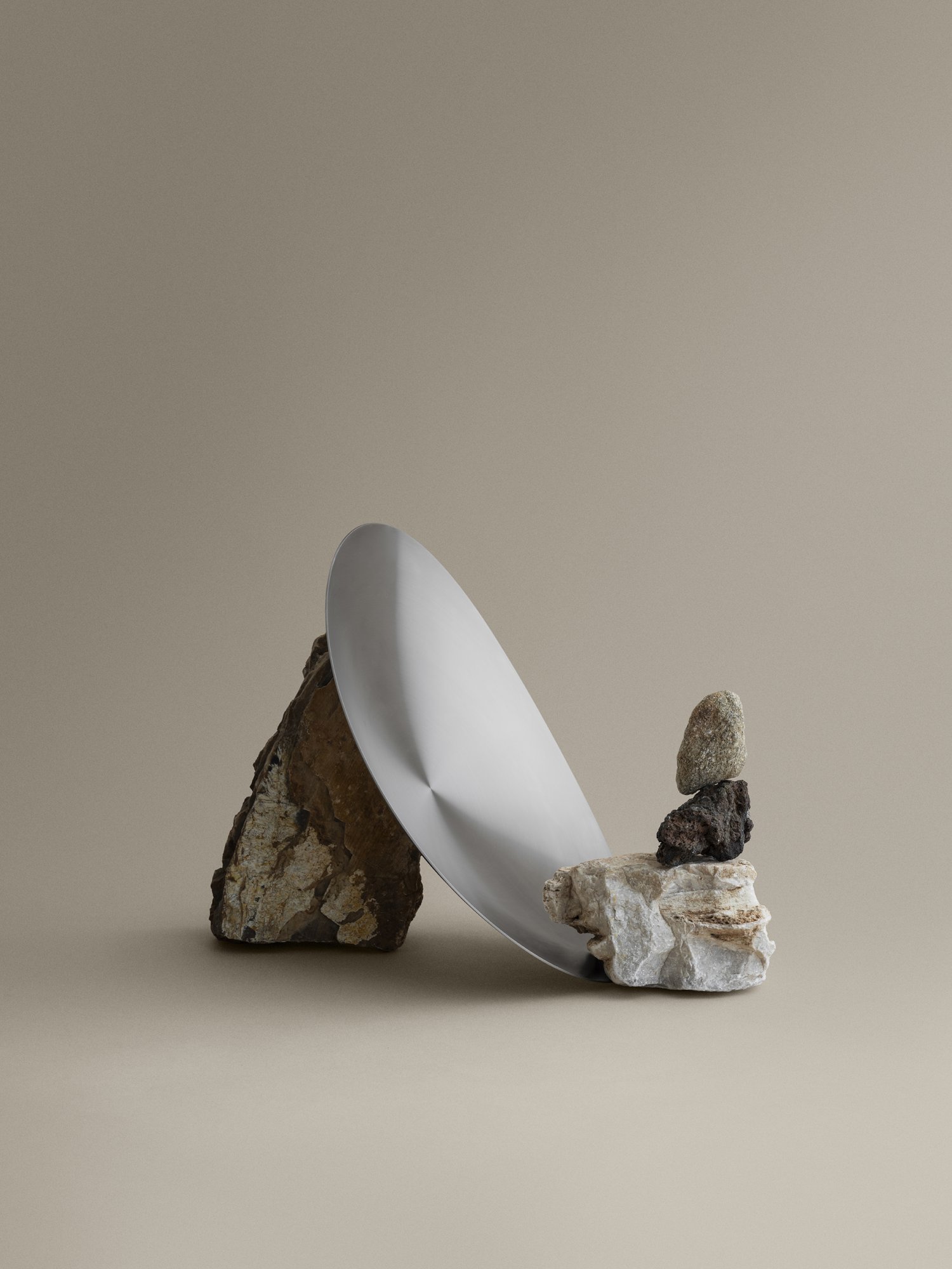 Minimalist still life featuring a metallic plate balanced on stacked rocks against a neutral backdrop.