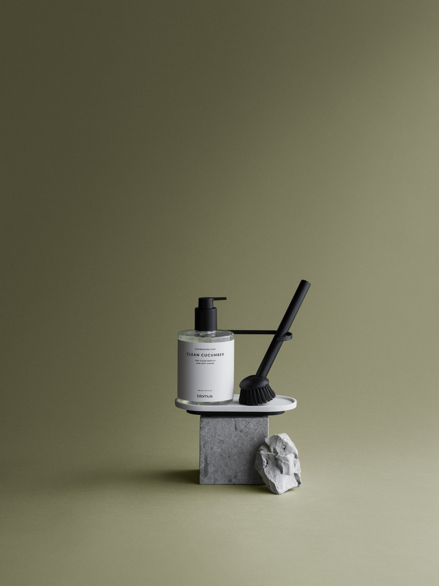 Minimalist hand soap and brush on a stone pedestal against a green background.