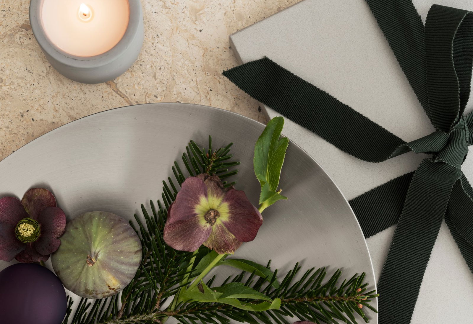 Elegant table setting with greenery, a flower, a fig, and a candle on a textured surface.