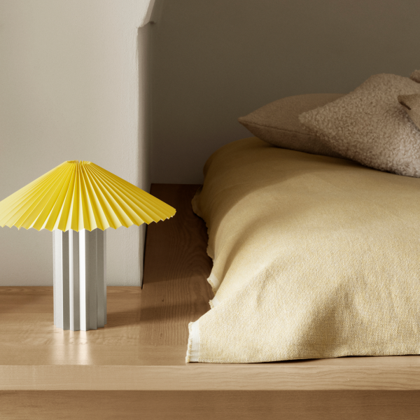 Moderne Nachttischlampe mit einem gelben Faltenschirm neben einem gemütlichen Bett mit weichen Kissen.