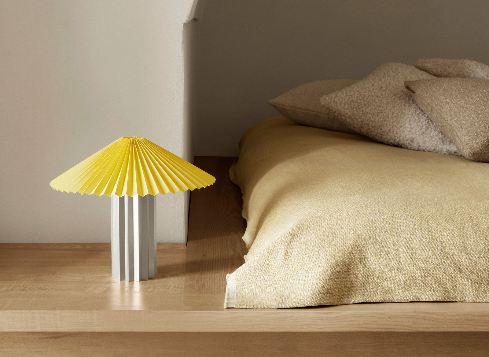 Moderne Nachttischlampe mit einem gelben Faltenschirm neben einem gemütlichen Bett mit weichen Kissen.