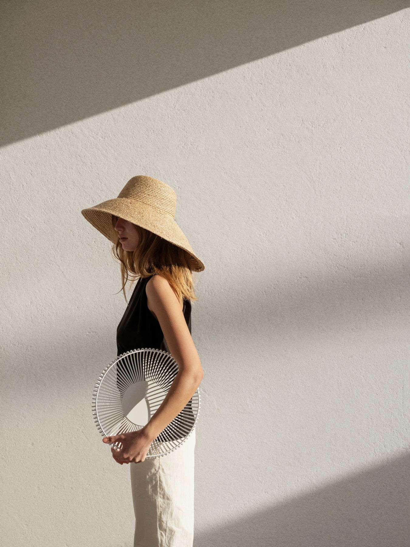 Woman in a straw hat holding a circular fan, standing against a minimalist backdrop.