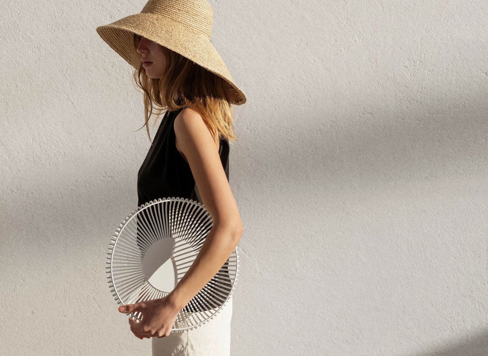 Woman wearing a straw hat holding a decorative white plate against a textured wall.