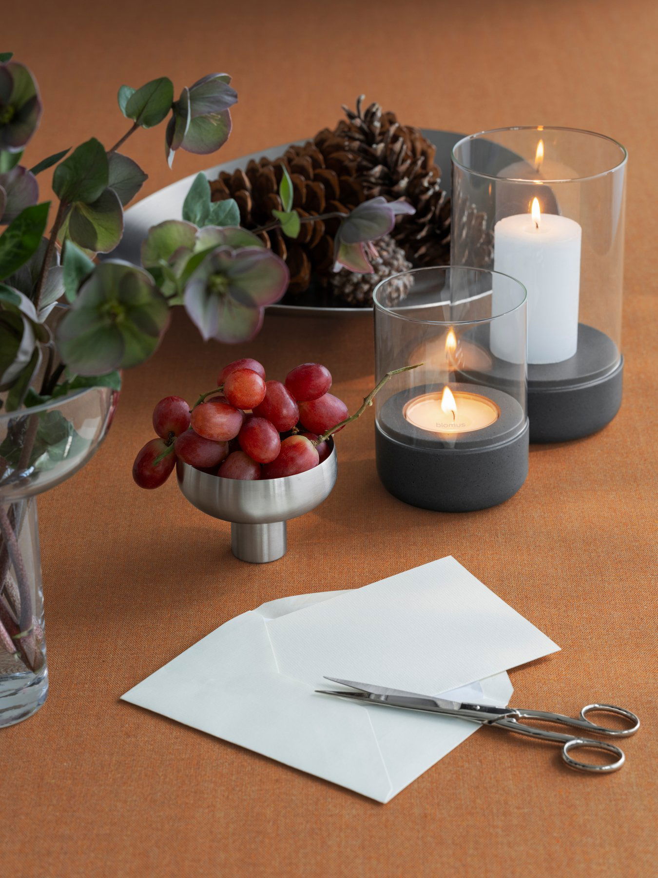 Stylische Tischdekoration mit Trauben, Kerzen und Tannenzapfen auf einer orangefarbenen Tischdecke.