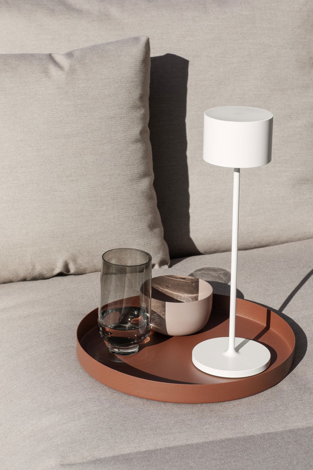 Modern lamp, glass, and bowl on a tray atop a stylish couch in natural light.