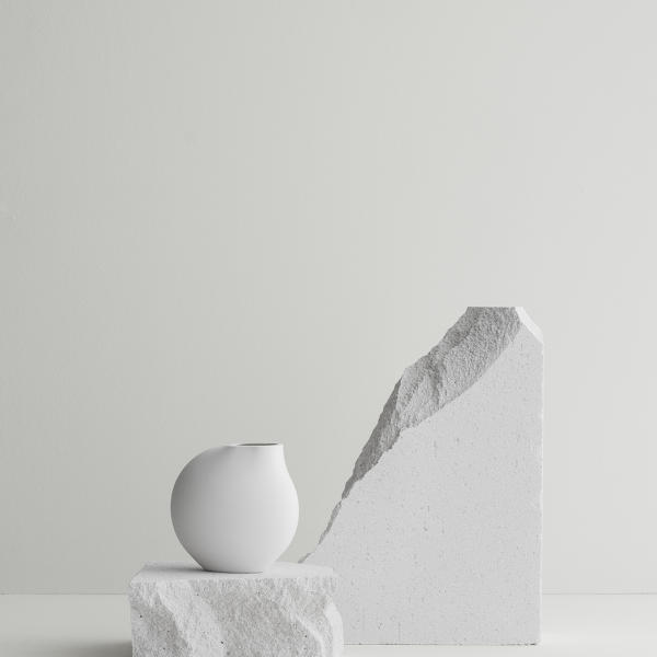 Minimalistische Komposition mit einer weißen Vase und strukturierten Steinblöcken vor einem hellen Hintergrund.
