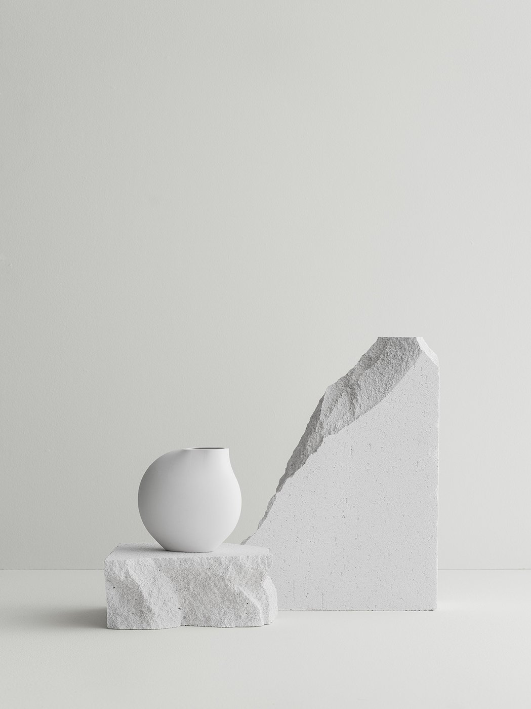 Minimalistische Komposition mit einer weißen Vase und strukturierten Steinblöcken vor einem hellen Hintergrund.