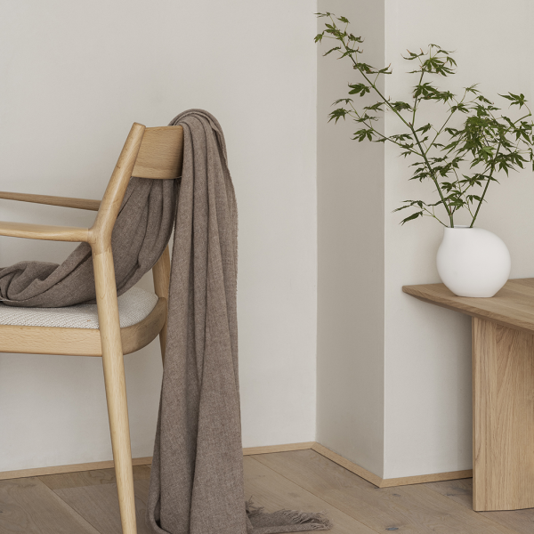 Gemütliches Interieur mit einem Stuhl, einer übergelegten Decke und einer Vase mit Grünpflanzen auf einem Holztisch.
