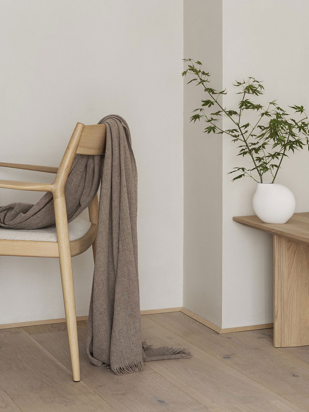 Gemütliches Interieur mit einem Stuhl, einer übergelegten Decke und einer Vase mit Grünpflanzen auf einem Holztisch.