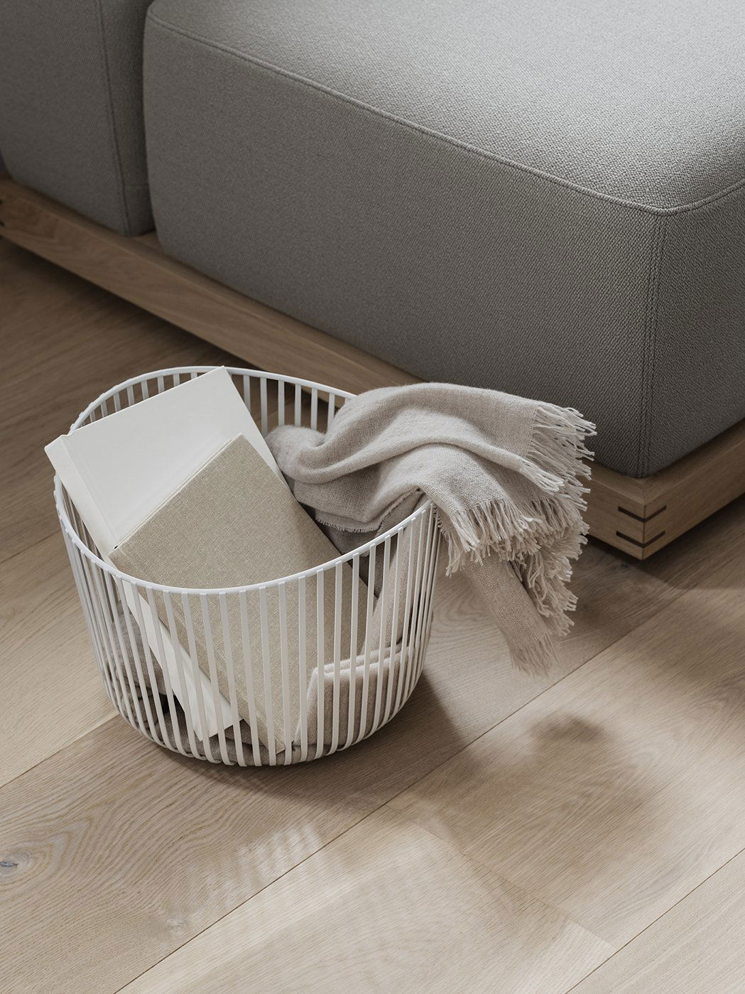 Weißer Drahtkorb mit gefaltetem Decken und Notizbüchern auf dem Holzboden neben einem Sofa.