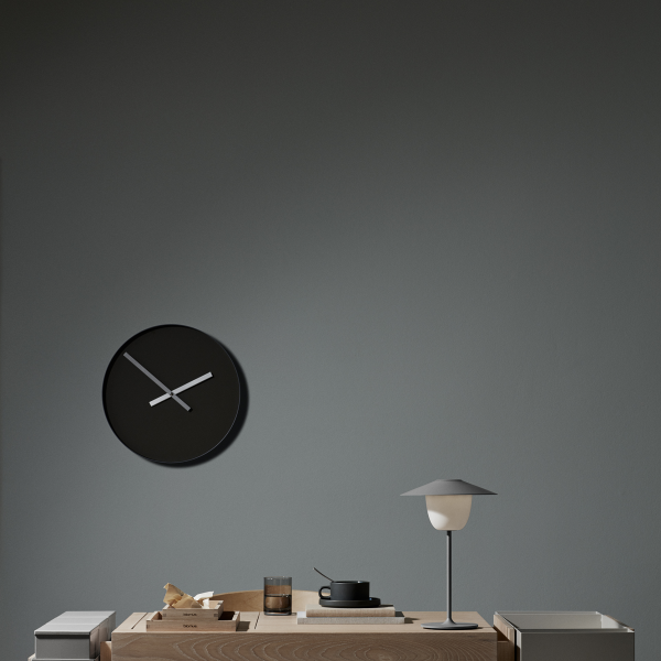 Minimalistische Innenraumszene mit einer modernen Uhr, Lampe und Holztisch vor einer grauen Wand.