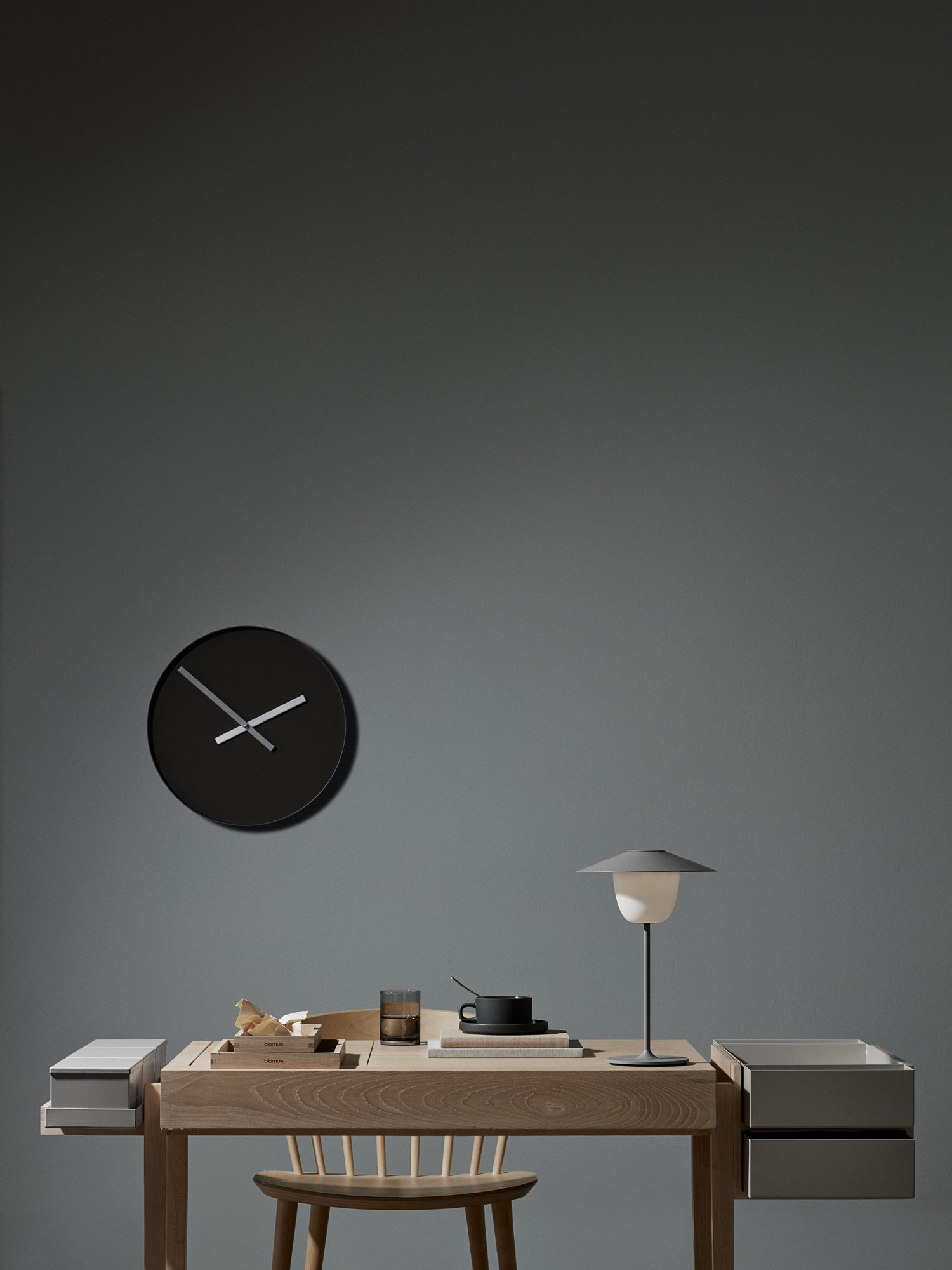 Minimalistische Innenraumszene mit einer modernen Uhr, Lampe und Holztisch vor einer grauen Wand.