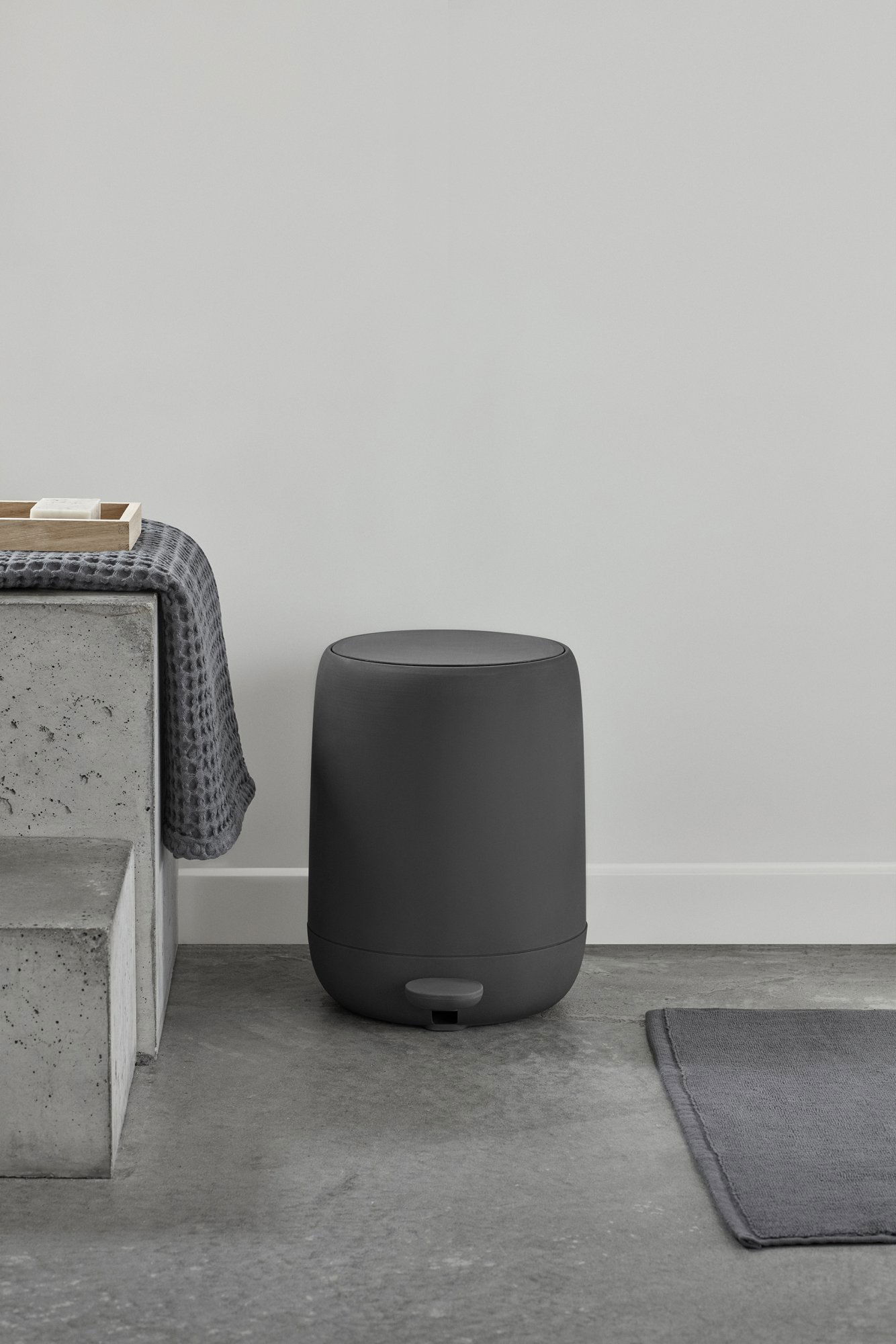 Modern black bathroom trash can beside a concrete bench in a minimalist setting.