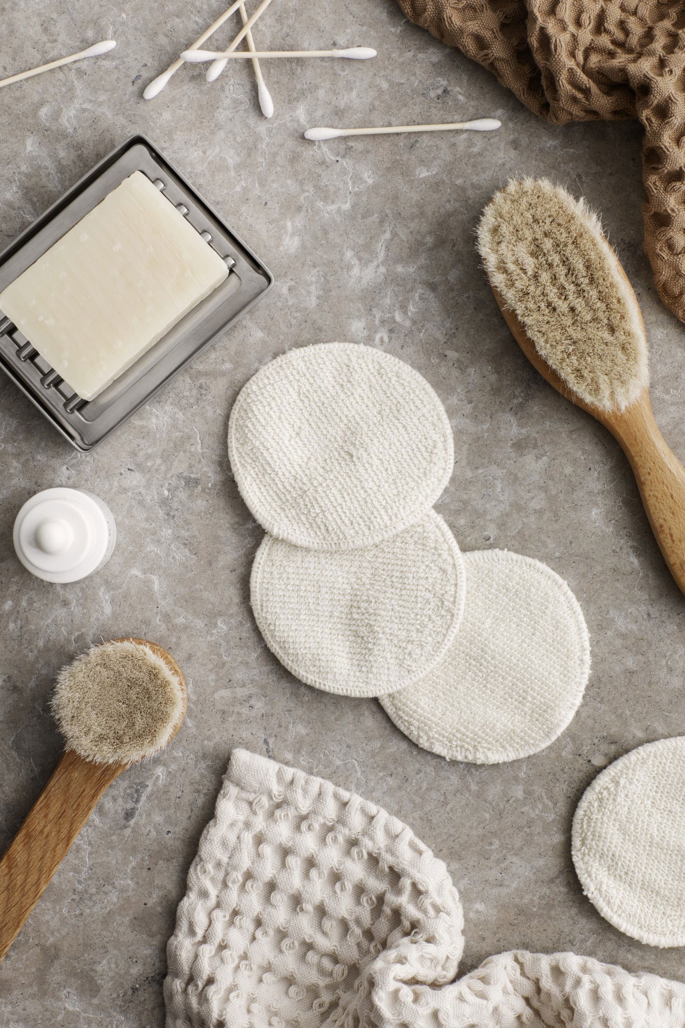 Natural skincare tools including reusable cotton pads, brushes, and soap on a textured surface.