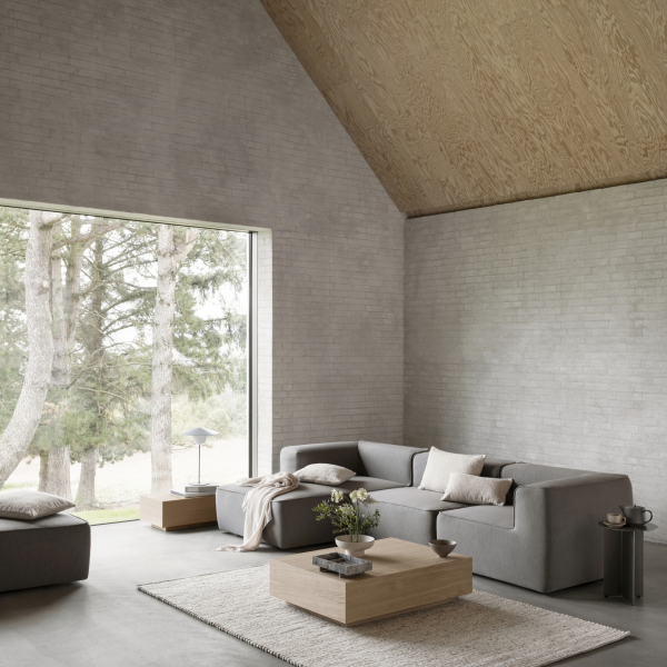 Moderner Wohnraum mit einem grauen Sofa, Holzakzenten und großen Fenstern, die die Natur zeigen.