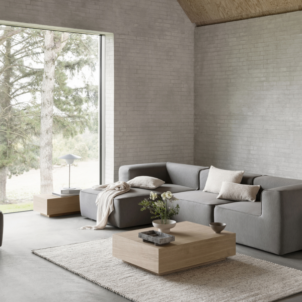 Moderner minimalistischer Wohnraum mit einem grauen Sofa und einem großen Fenster, das die Außenanlagen mit Grünpflanzen zeigt.