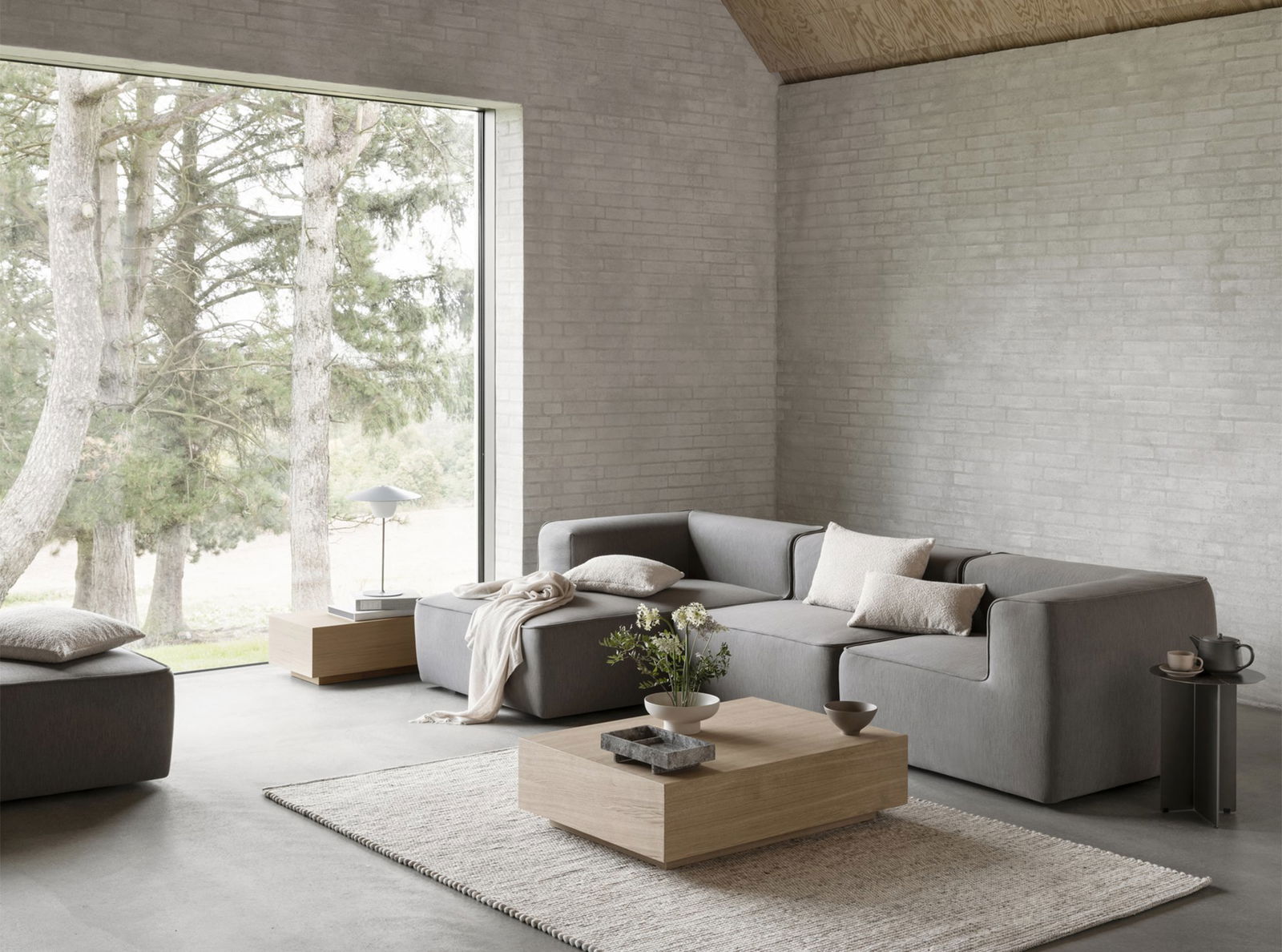 Moderner minimalistischer Wohnraum mit einem grauen Sofa und einem großen Fenster, das die Außenanlagen mit Grünpflanzen zeigt.