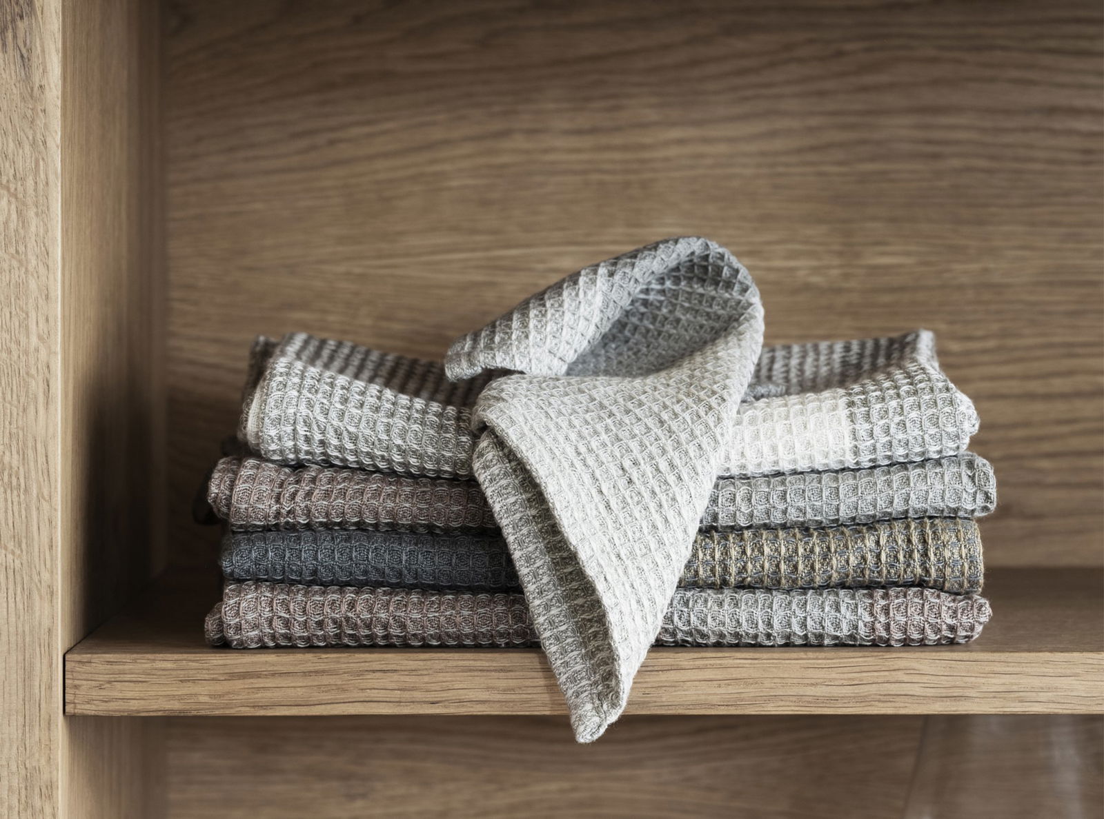 Stack of textured kitchen towels in neutral colors displayed on a wooden shelf.