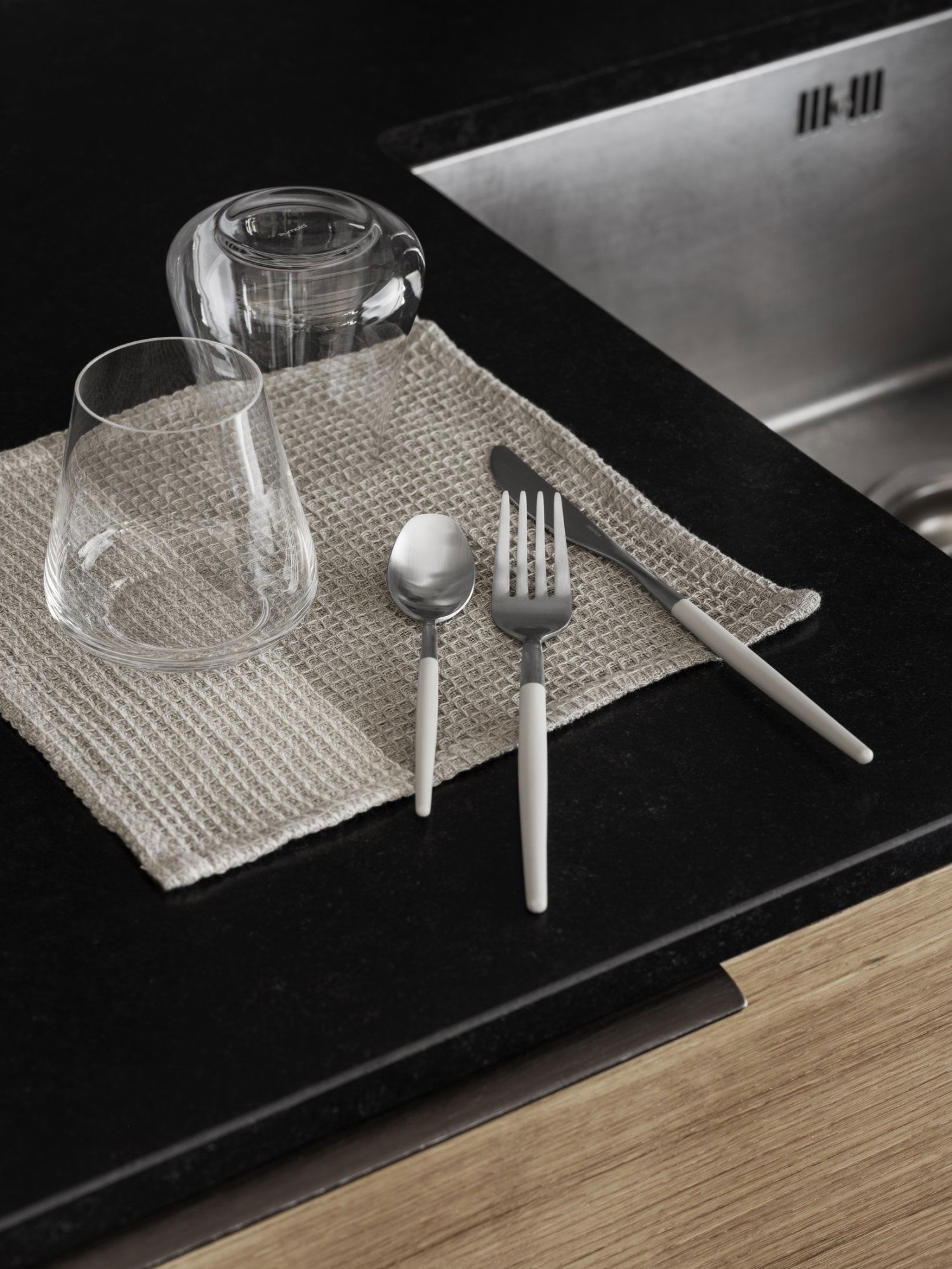 Modern dining setup with cutlery, glassware, and a textured placemat on a dark countertop.