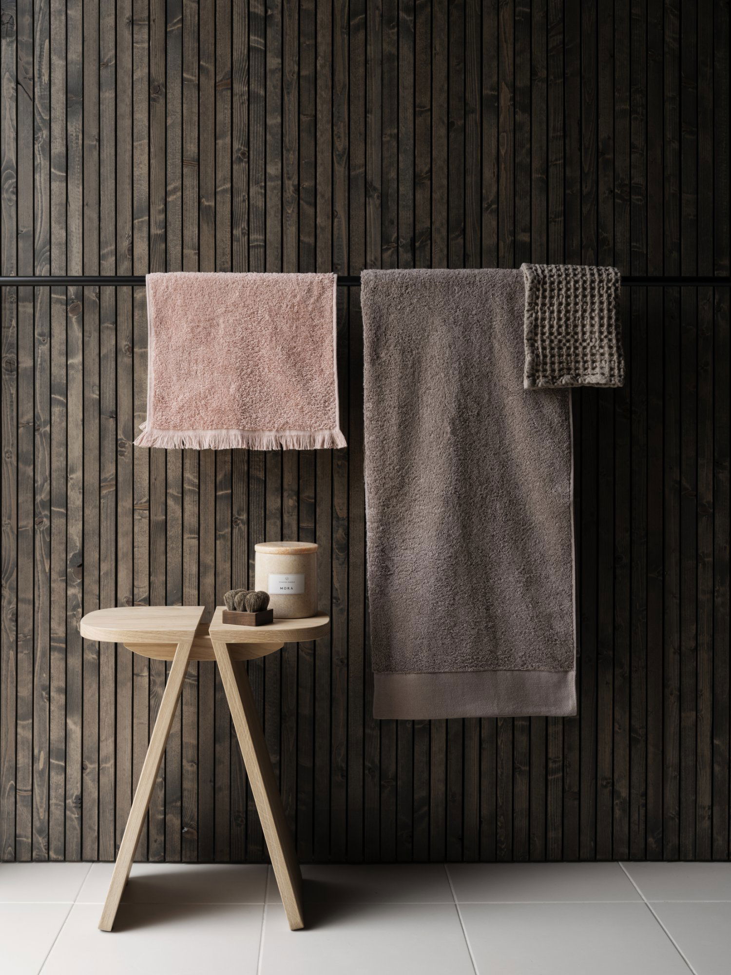 Soft pink and grey towels hanging on a rustic wooden rack next to a candle on a side table.