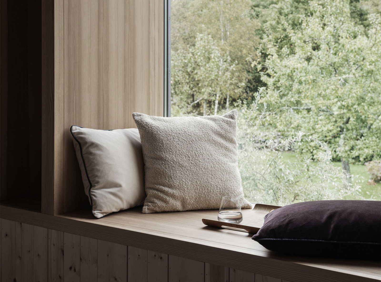 Gemütlicher Fenstersitz mit weichen Kissen und einem Glas auf einem Holzregal mit Blick auf die Natur.