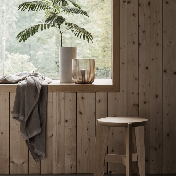 Cozy indoor scene with a plant, vase, throw blanket, and wooden stool by a large window.