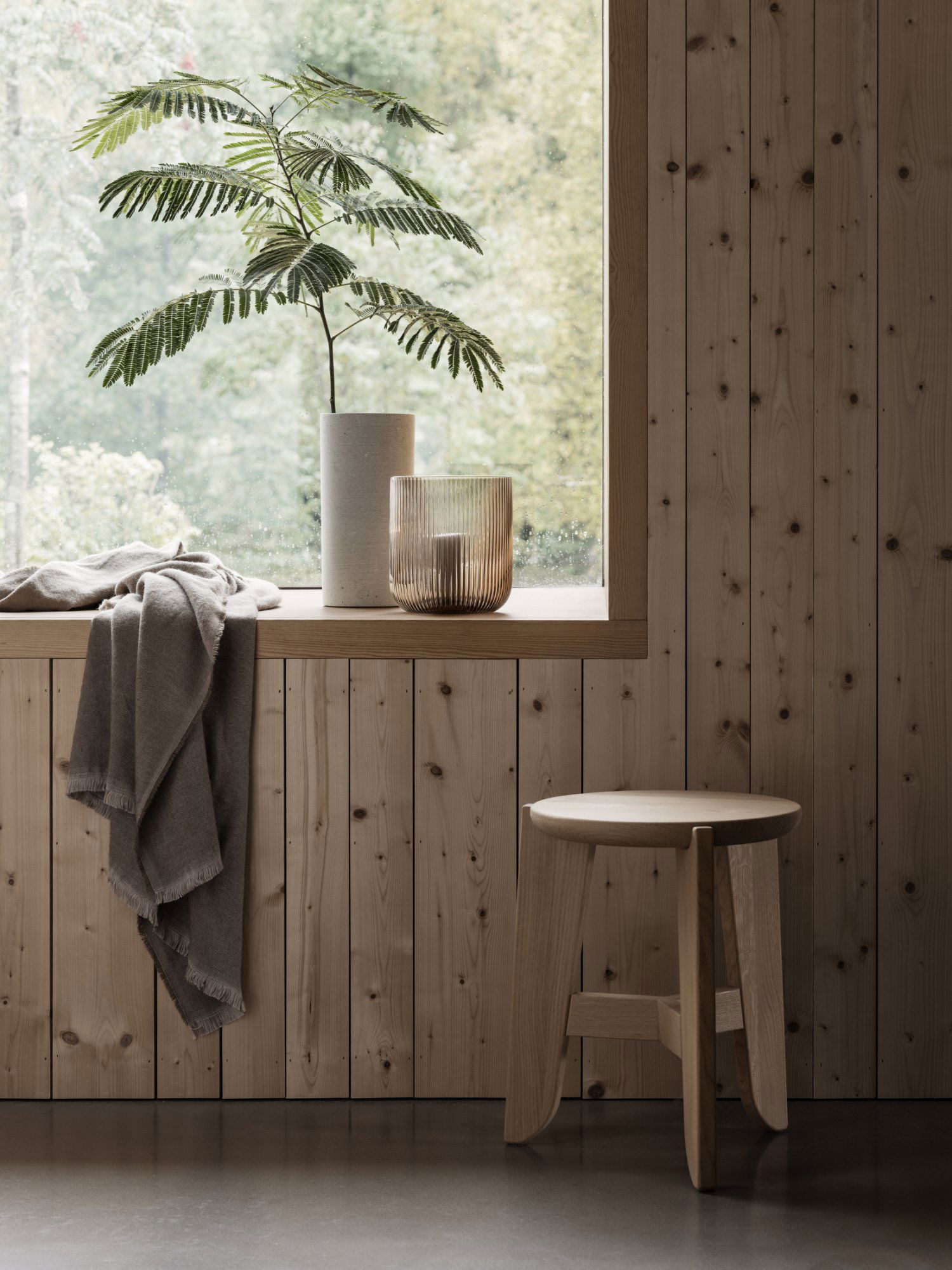 Cozy indoor scene with a plant, vase, throw blanket, and wooden stool by a large window.