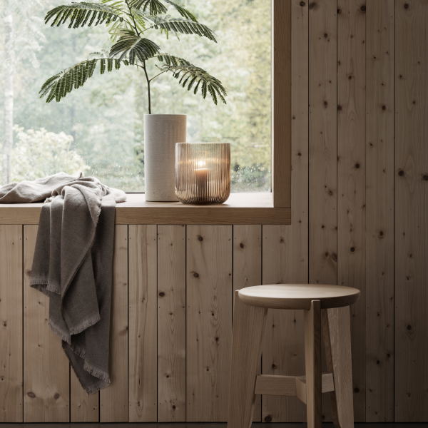 Cozy interior with wooden walls, a plant, candle holder, and a simple stool by the window.