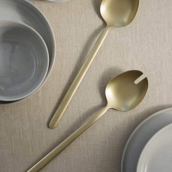 Elegant gold serving utensils beside gray bowls on a textured linen tablecloth.