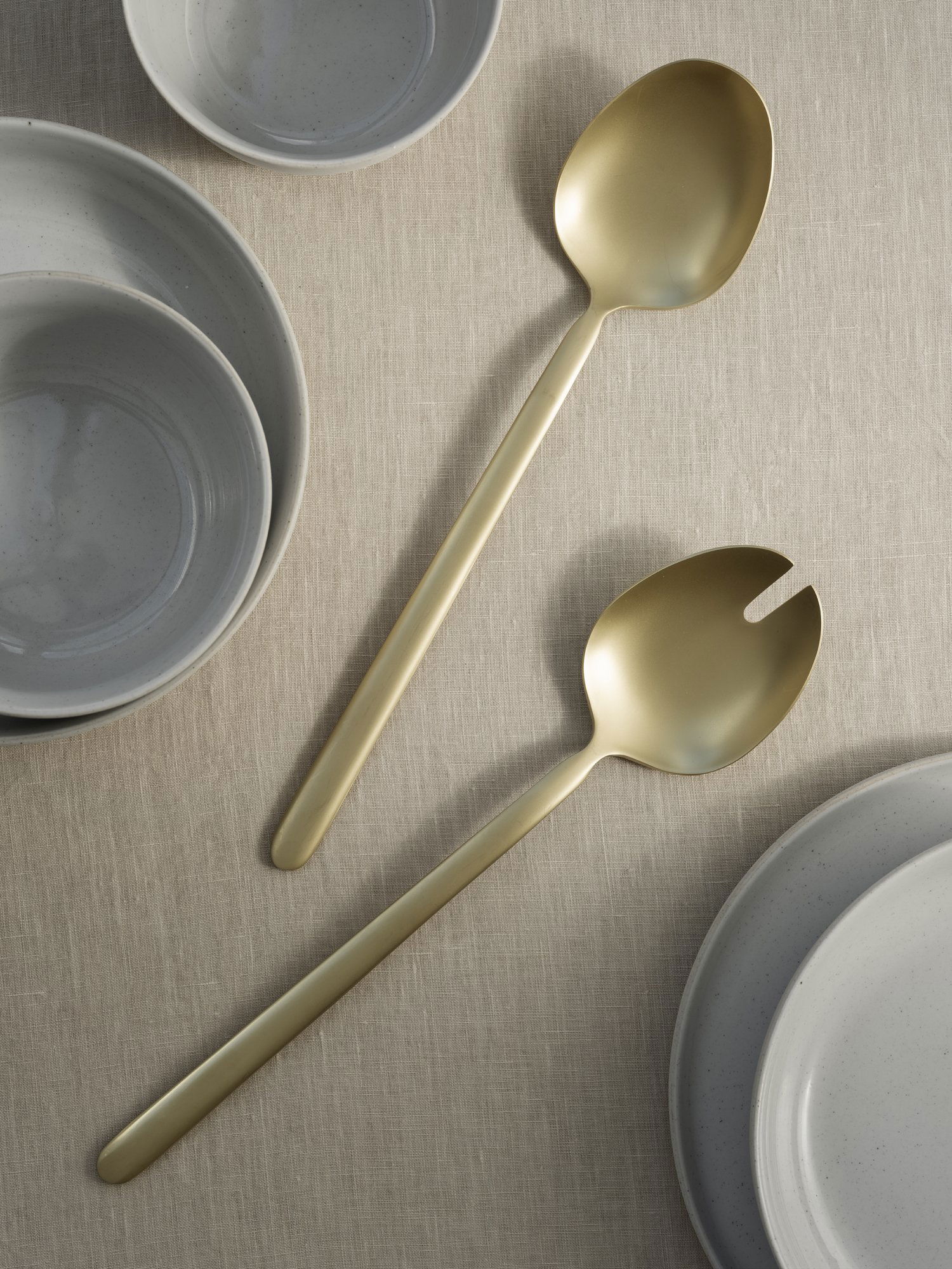 Elegant gold serving utensils beside gray bowls on a textured linen tablecloth.