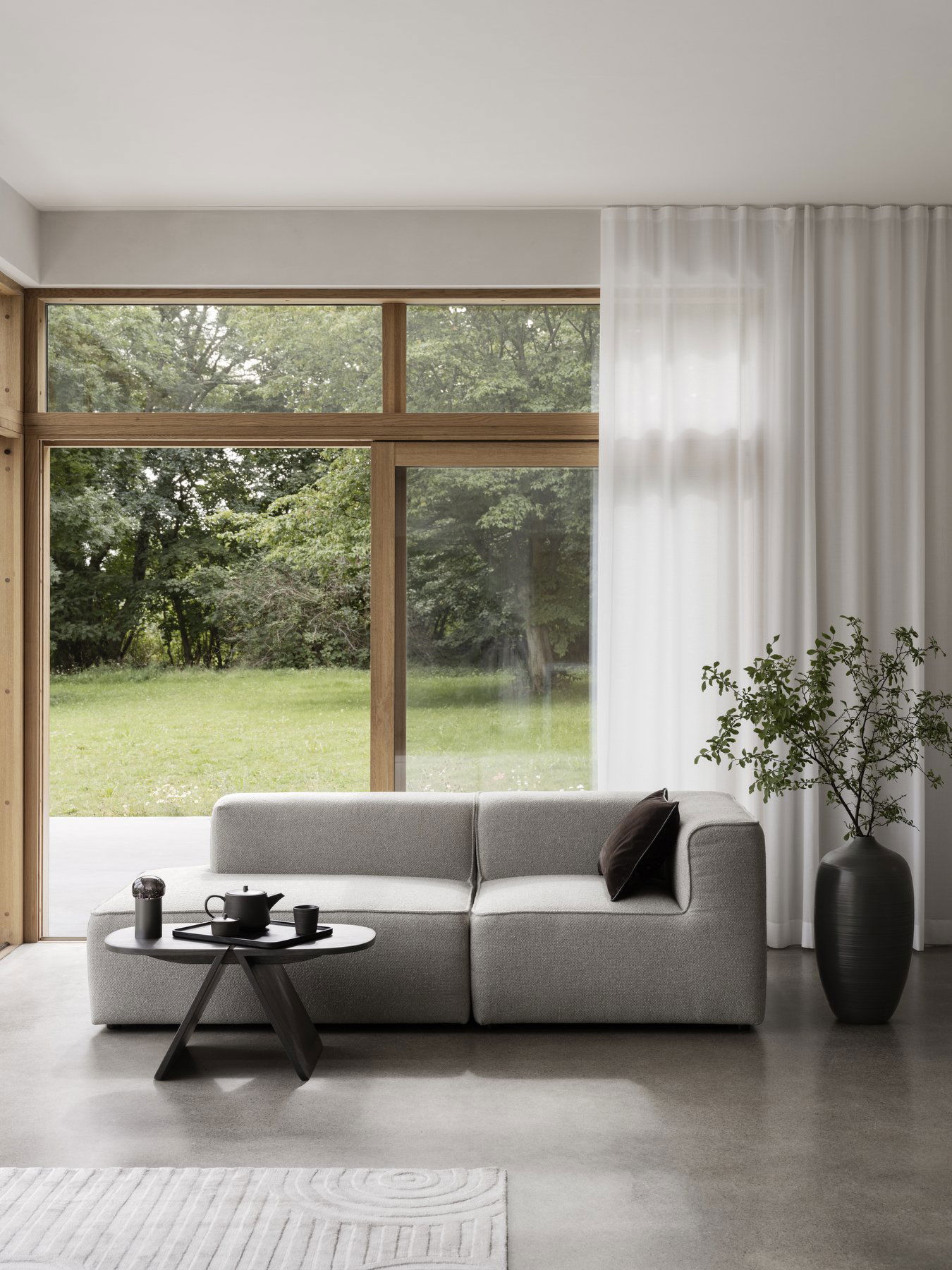Stylisches modernes Wohnzimmer mit grauem Sofa, Kaffeetisch und großen Fenstern mit Blick auf das Grüne.