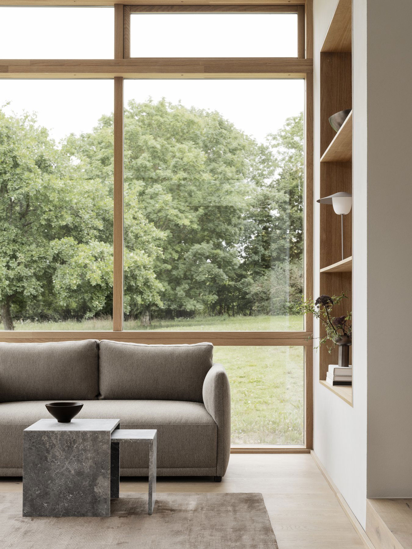 Modern living room with a gray sofa, stylish decor, and large windows overlooking greenery.