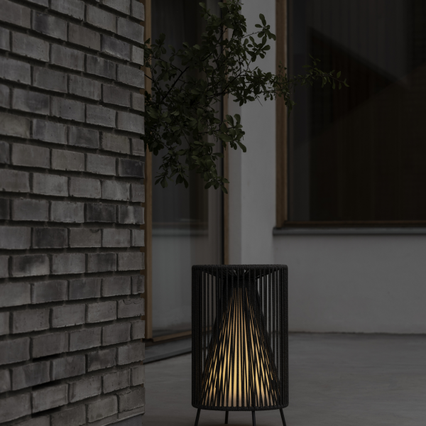 Moderne schwarze Lampe mit leuchtendem Innenraum, platziert neben einer strukturierten Backsteinmauer.