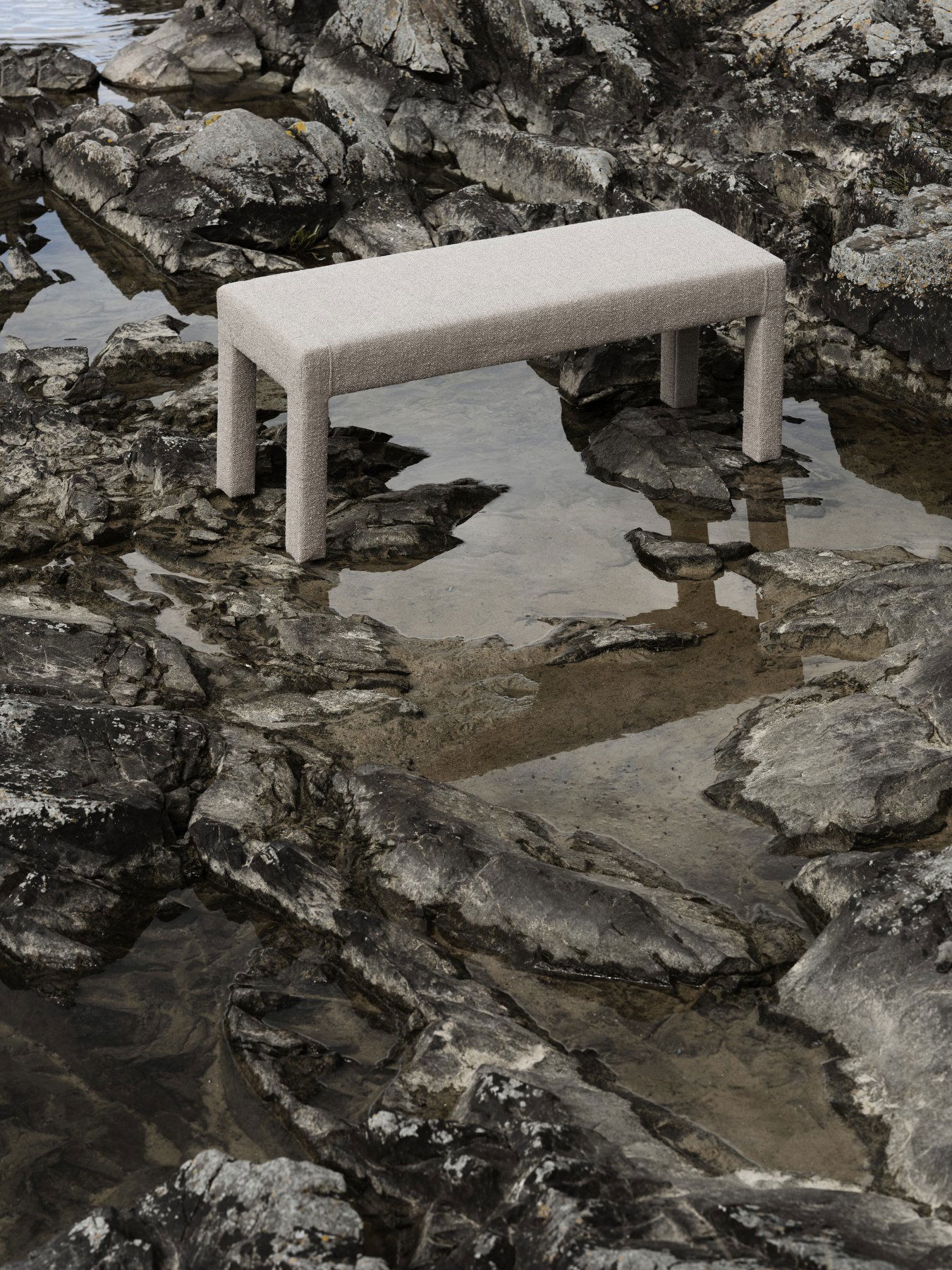 Eine stilvolle beige Bank auf felsigem Gelände, die sich im ruhigen Wasser spiegelt.